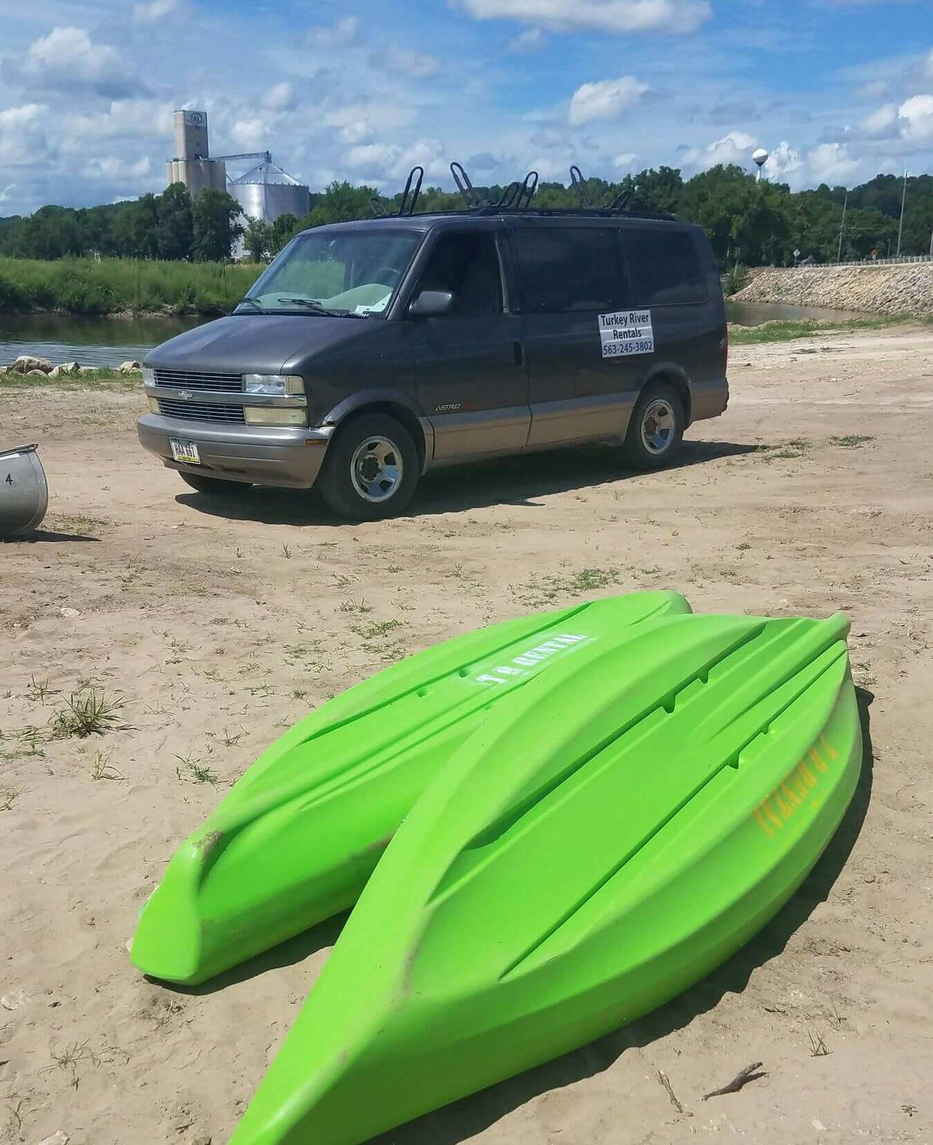 Two green kayaks on sand with a gray van parked in the background near a river.
