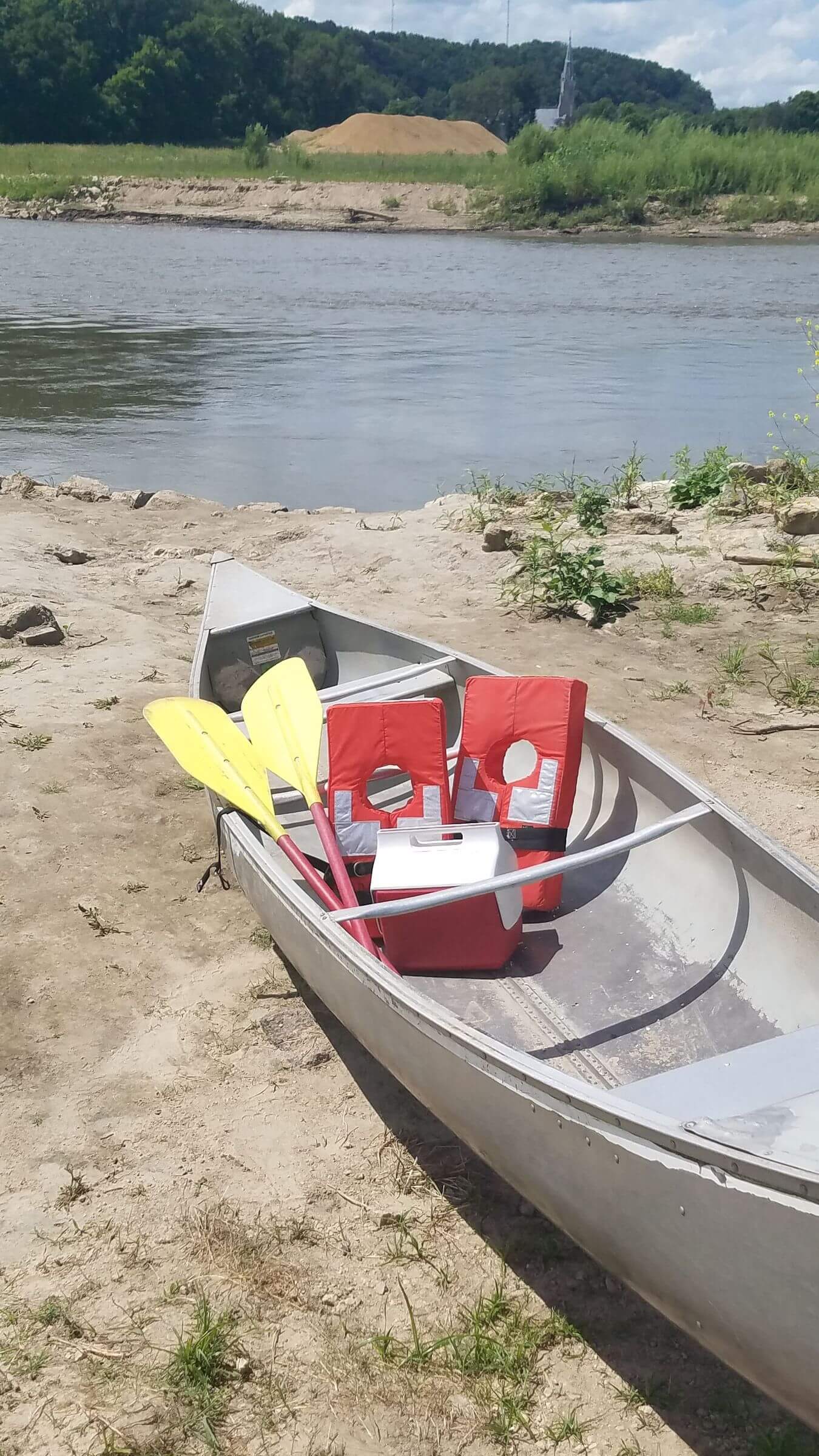 A canoe with paddles, chairs, and a cooler on a sandy riverbank under a sunny sky.