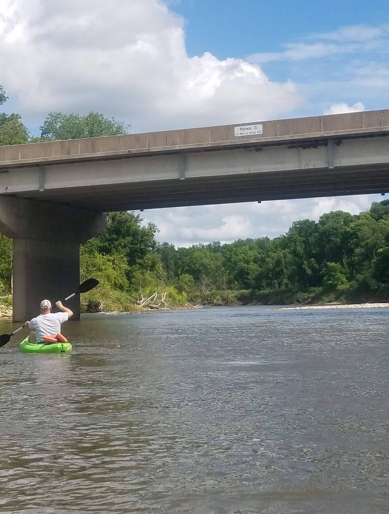 Person kayaking under a concrete bridge on a calm river with trees and clouds in the background.
