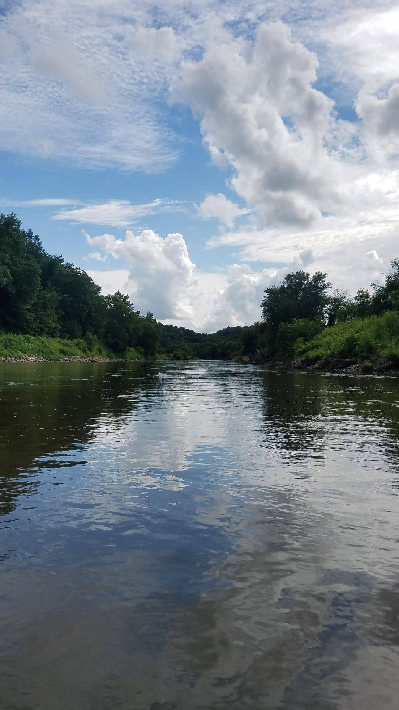 A calm river flows between green trees under a blue sky with scattered clouds.