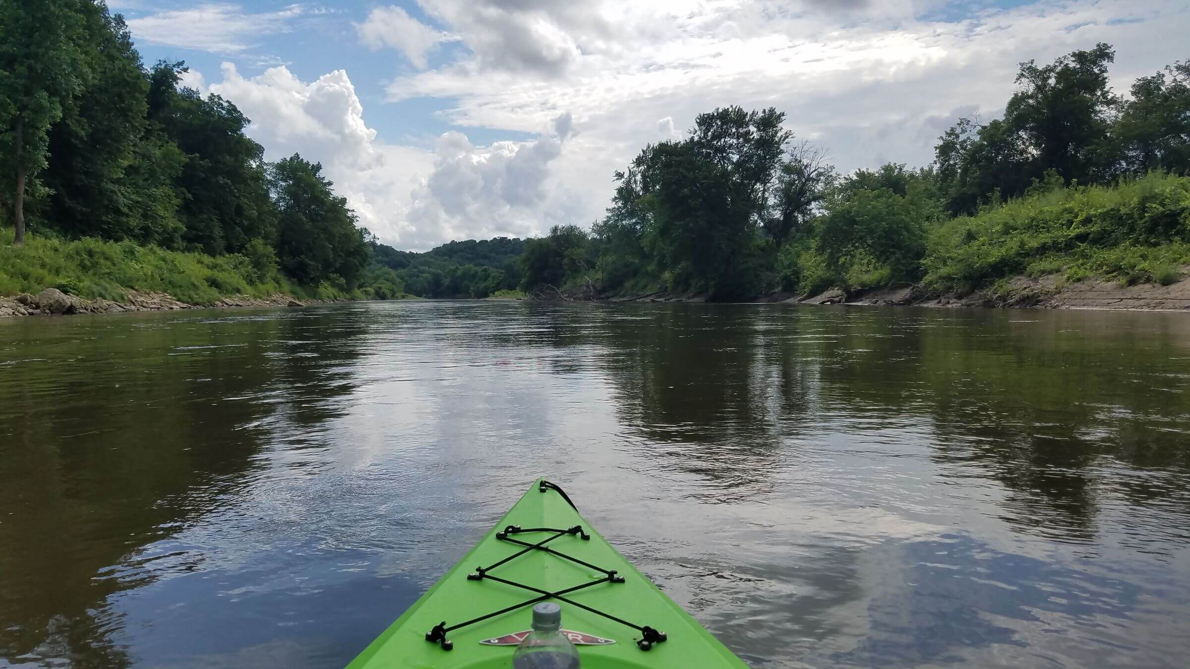 Green kayak on a calm river surrounded by trees under a partly cloudy sky.